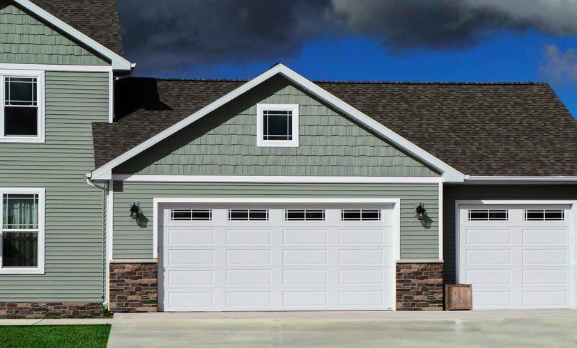 Two-story house with green siding, white garage doors, and brown stone accents against a blue sky.