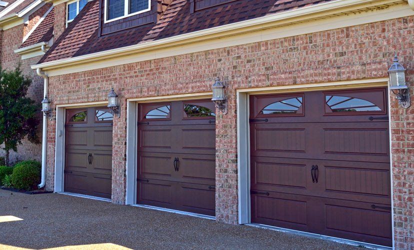 Three brown garage doors on a brick building, with arched windows and decorative lighting.