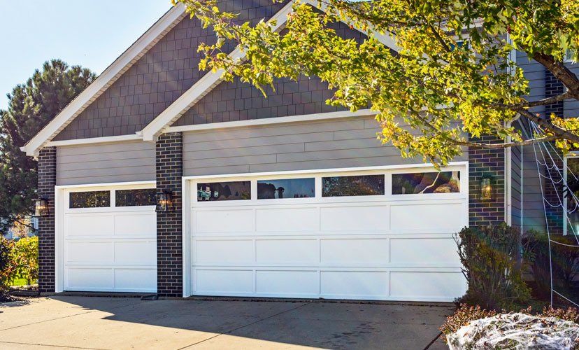 White garage doors on a house with gray siding, brick pillars, and a tree in the foreground.