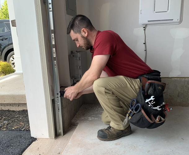 Services Man in red shirt, khaki pants, and work boots works on a garage door. He is crouched and using tools.
