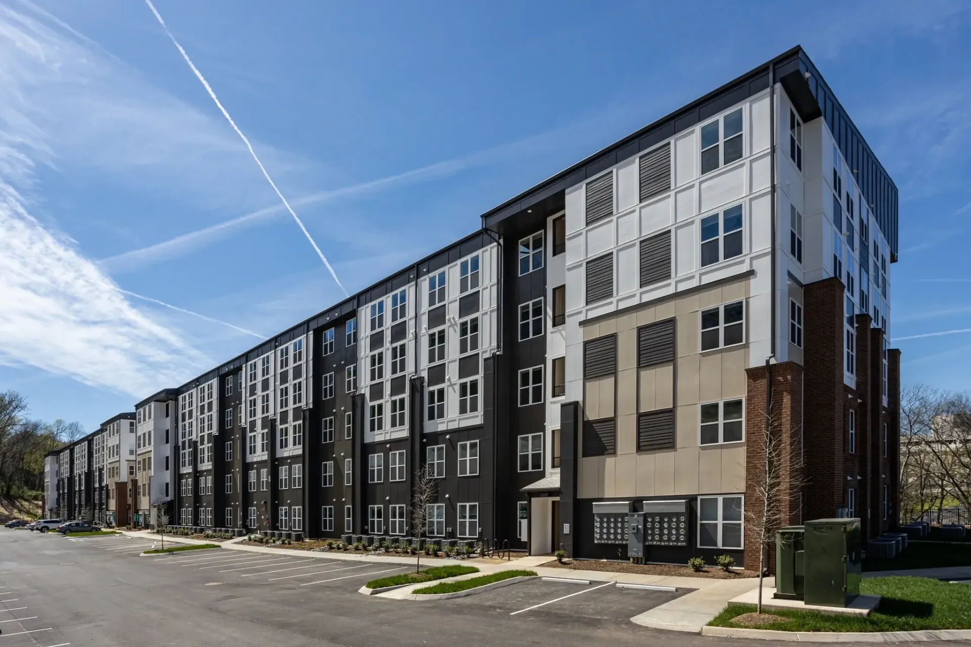 Exterior view of a modern apartment building with a parking lot and blue sky with contrails.