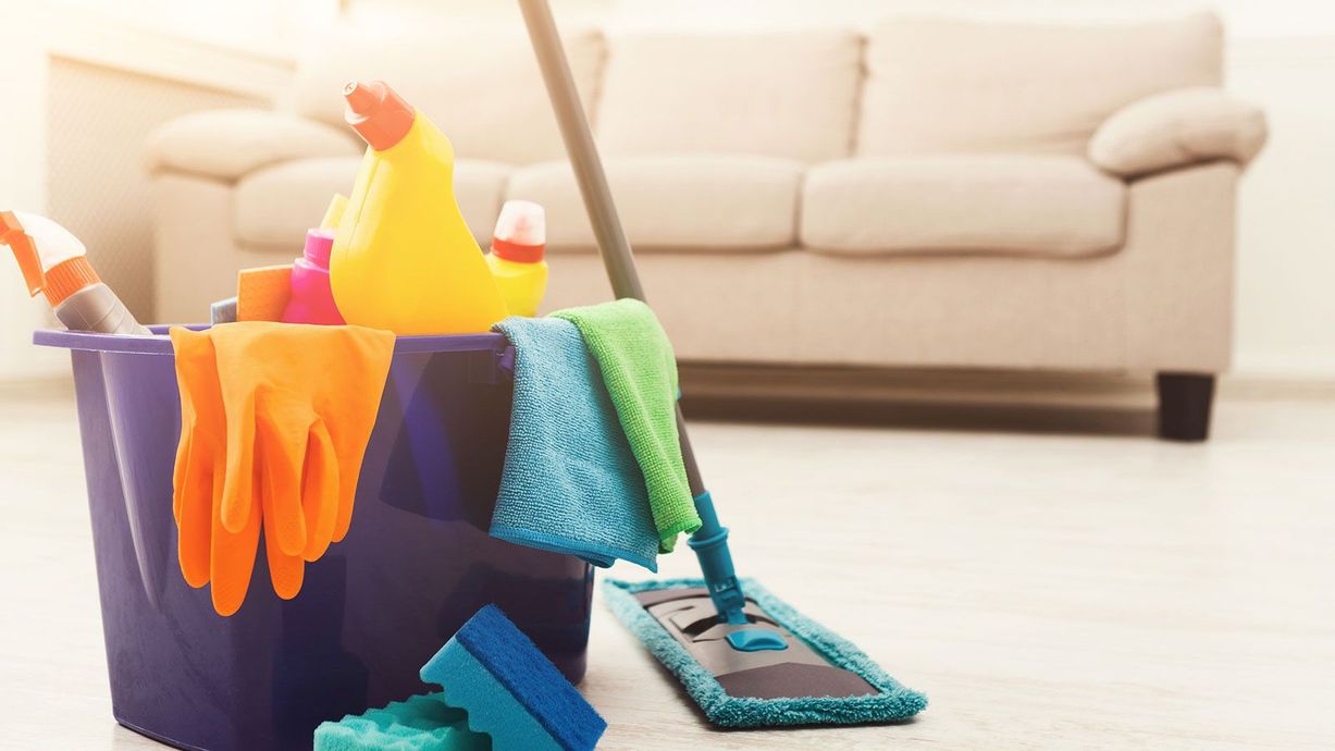 Colorful cleaning supplies in a bucket with rubber gloves and a mop on a bright living room floor