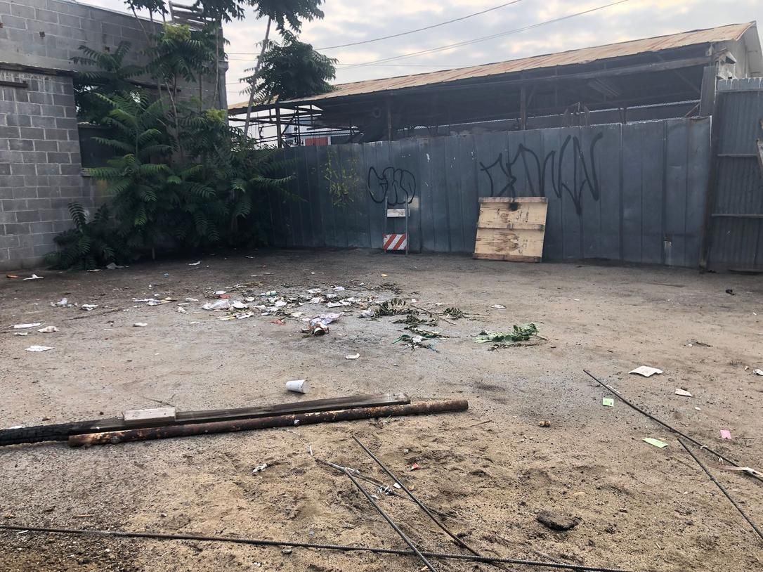 Dirt lot beside a corrugated metal gate, with scattered debris and a small tree on the left.