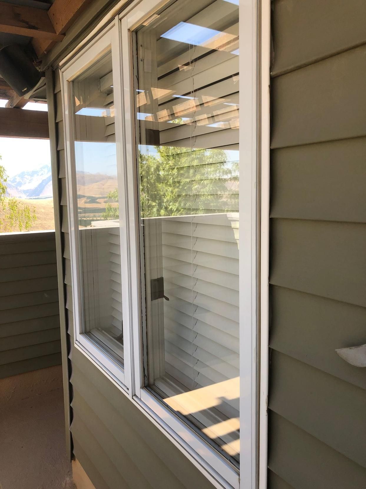 Exterior view of a house window with white trim and beige siding, reflecting trees and sky