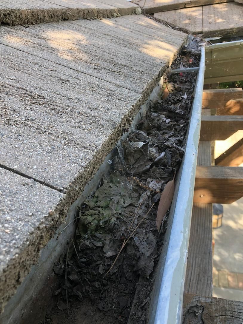 Gutter filled with leaves beside a concrete sidewalk and wooden deck railing.