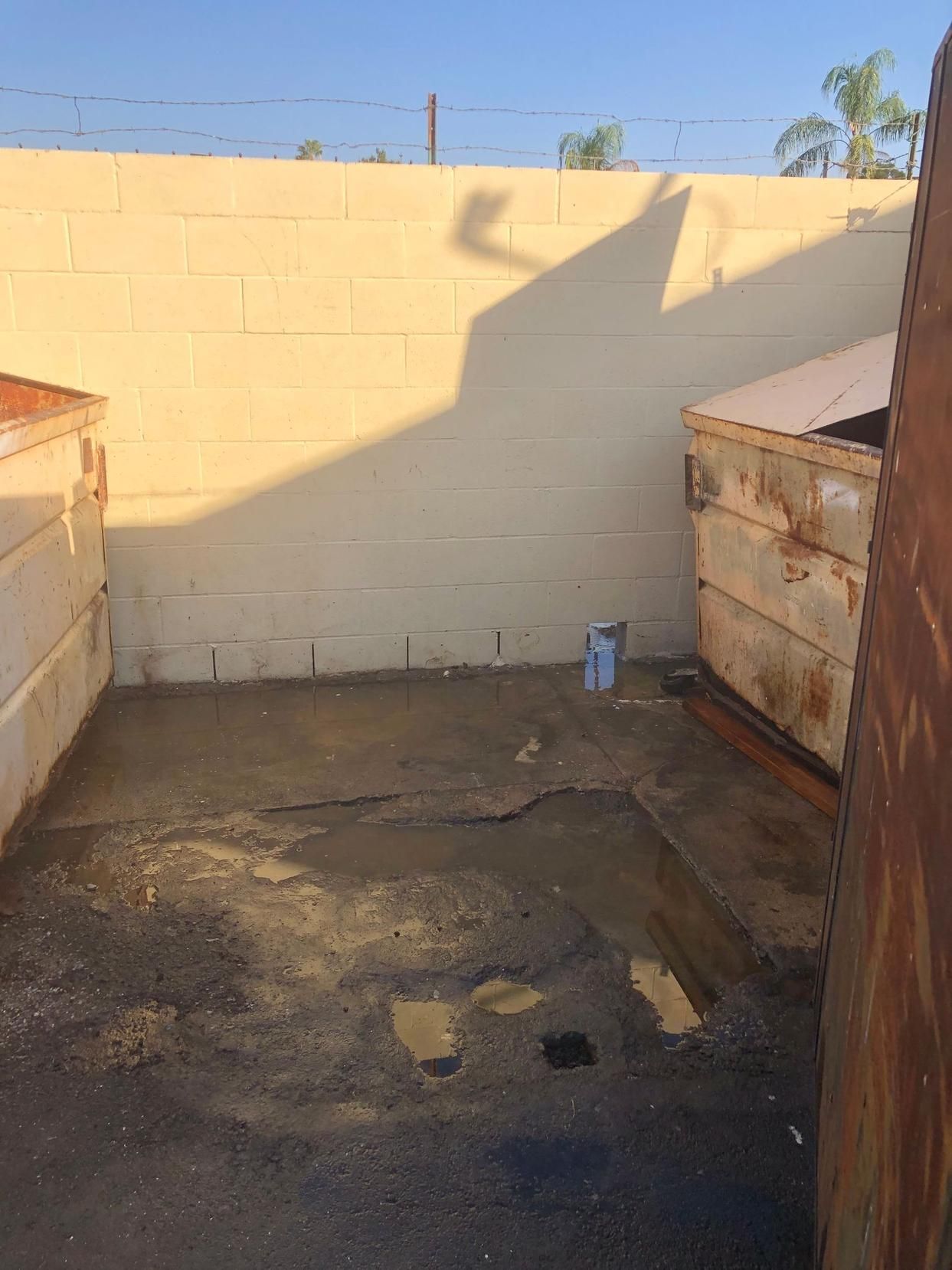 Sunlit alley between rusty dumpsters and a beige wall, with a stained concrete ground.