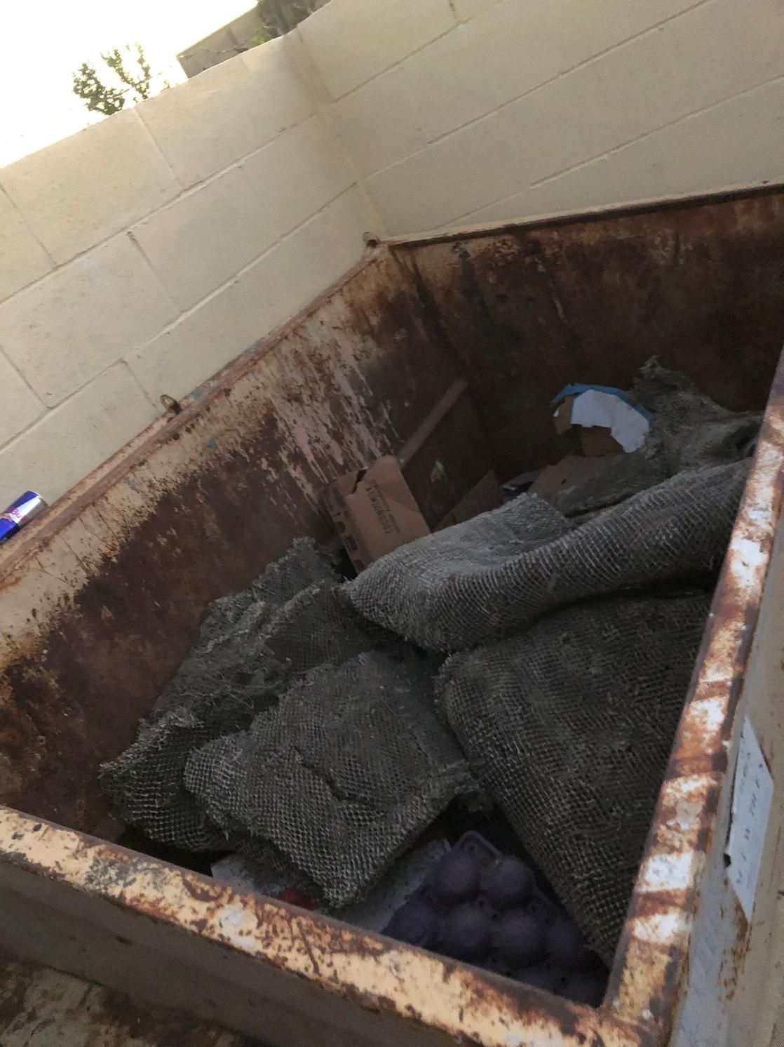 Rusty dumpster filled with folded black carpet or turf pieces outdoors
