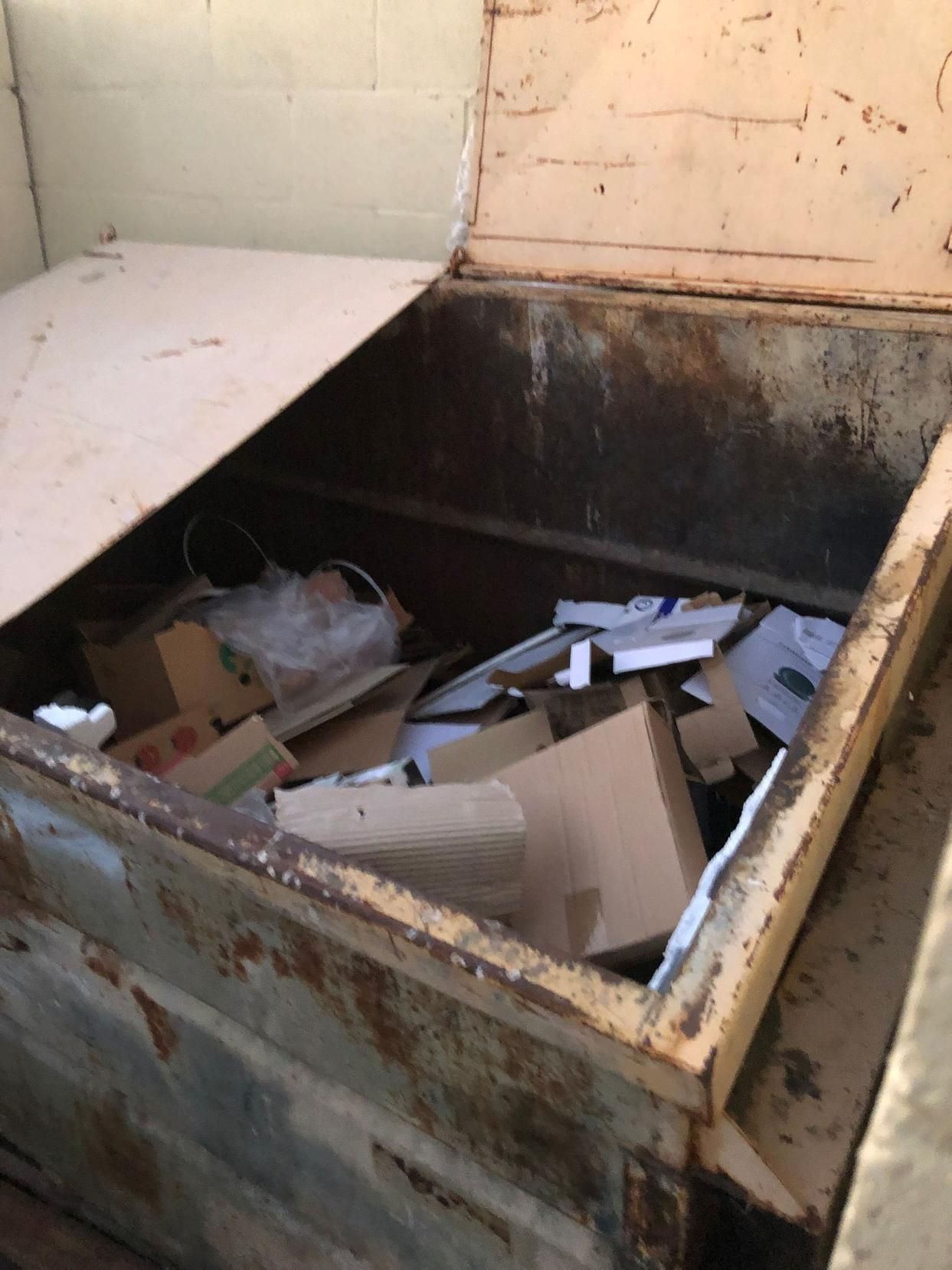 Open dumpster with scattered cardboard and trash inside, beside a weathered wall.