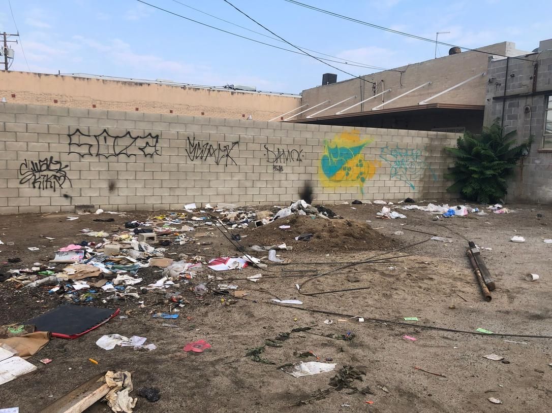 Trash-strewn dirt lot beside a graffiti-marked concrete wall under a clear sky.