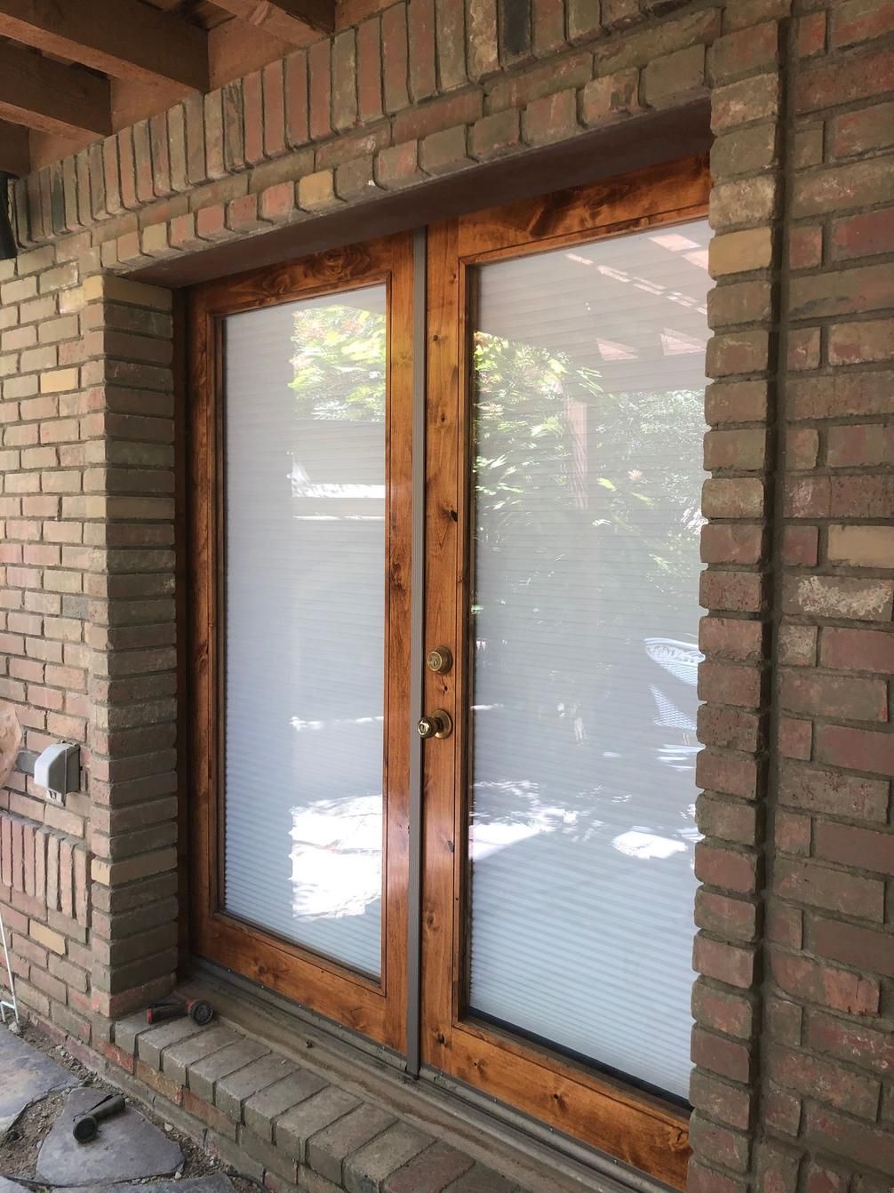 Wooden-framed glass double doors set in a brick wall, with frosted lower panels and sunlight reflections.