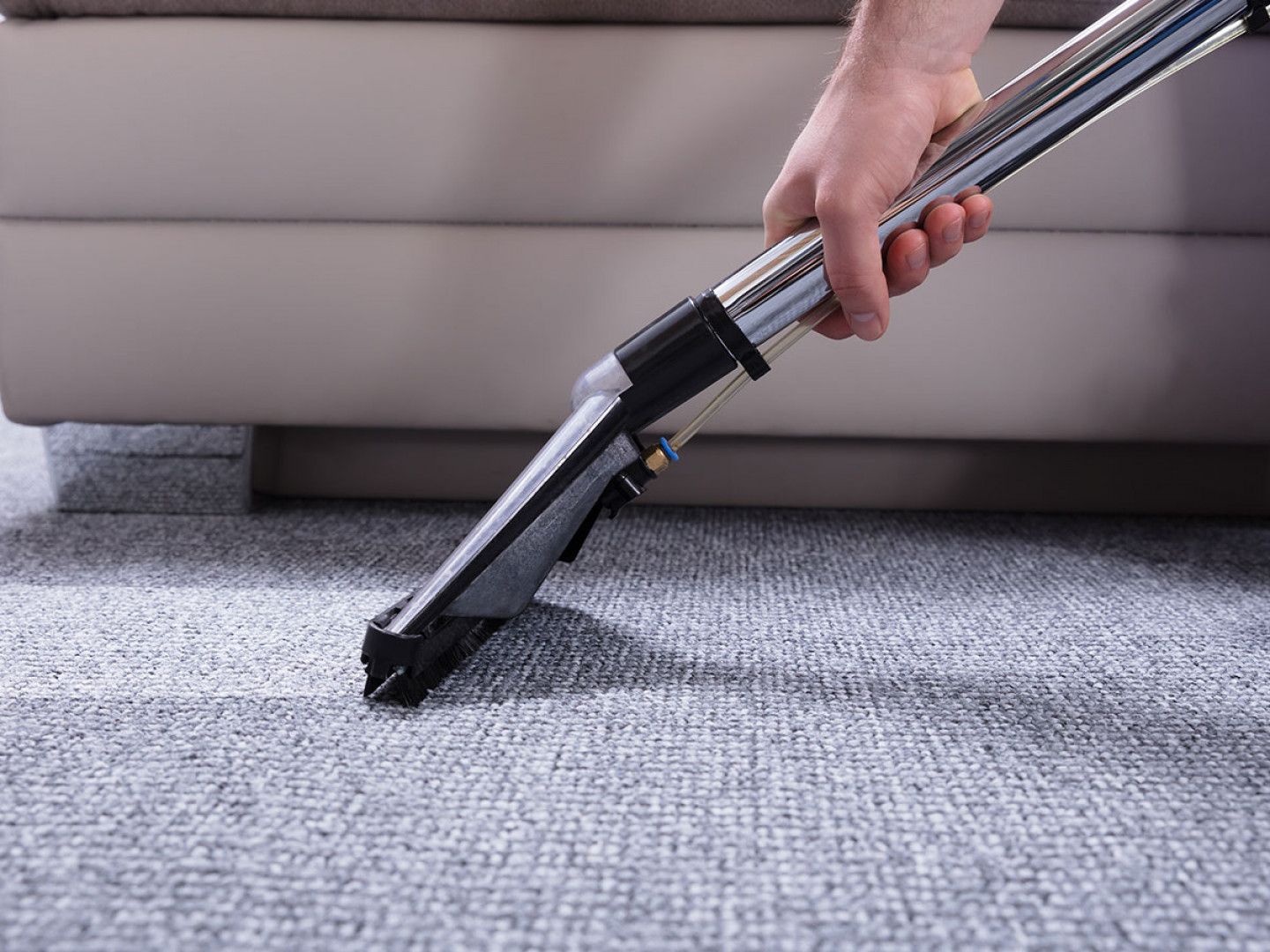Hand using a carpet cleaner nozzle on gray carpet beside a beige sofa