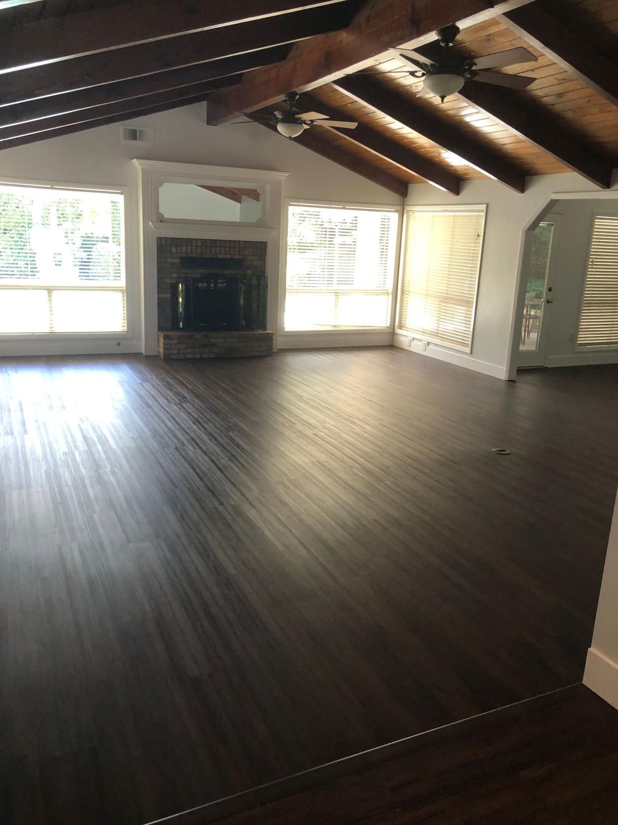 Empty living room with dark wood floors, exposed ceiling beams, fireplace, and large windows.