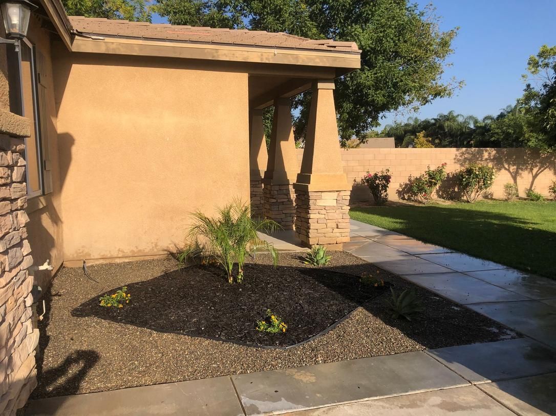 Front yard with tan stucco house, stone porch, mulch bed, and green lawn in sunlight