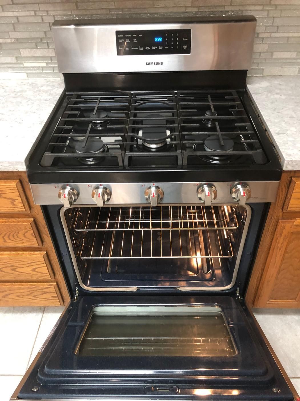 Stainless steel gas stove with oven door open, showing racks and lit control panel in a kitchen.
