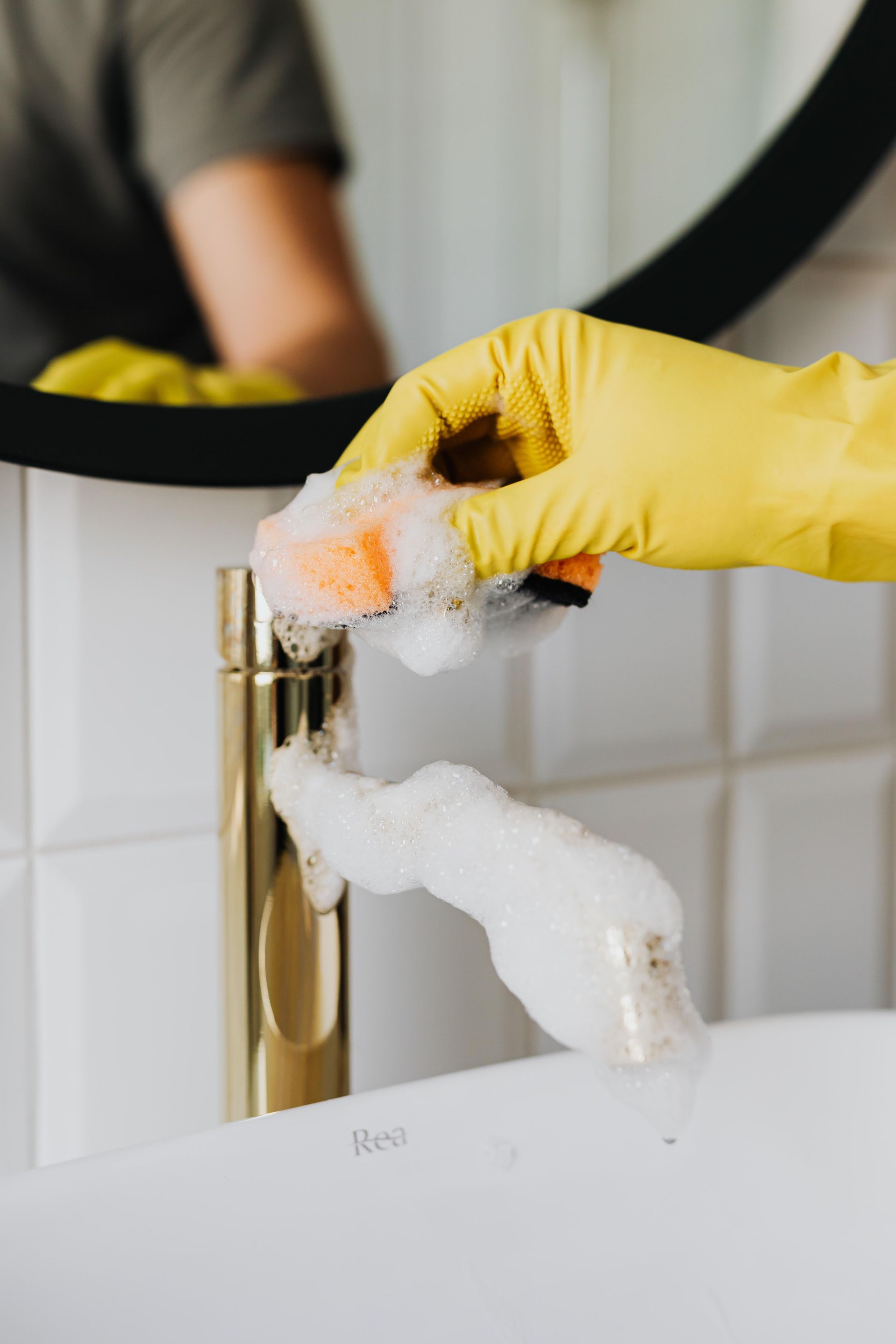 Yellow-gloved hand scrubbing foam from a gold faucet at a bathroom sink
