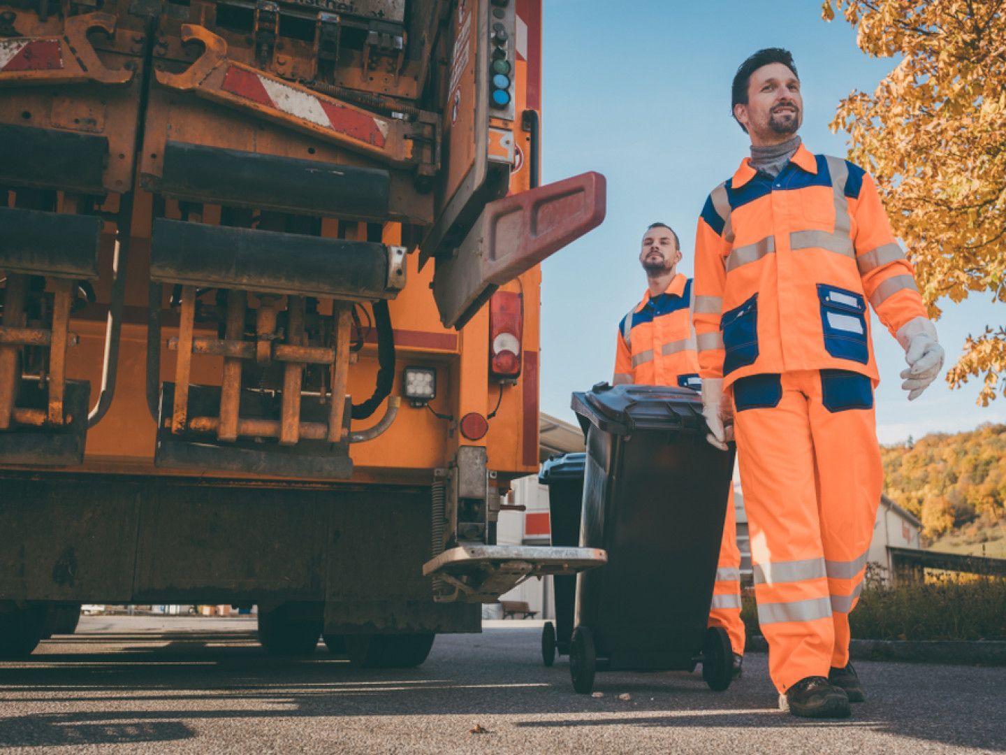 Workers in orange uniforms beside a garbage truck, rolling bins on a street in autumn.