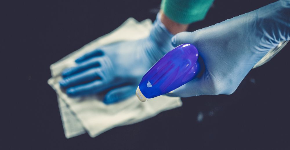 Gloved hands wiping a surface with a cloth, one holding a blue spray bottle