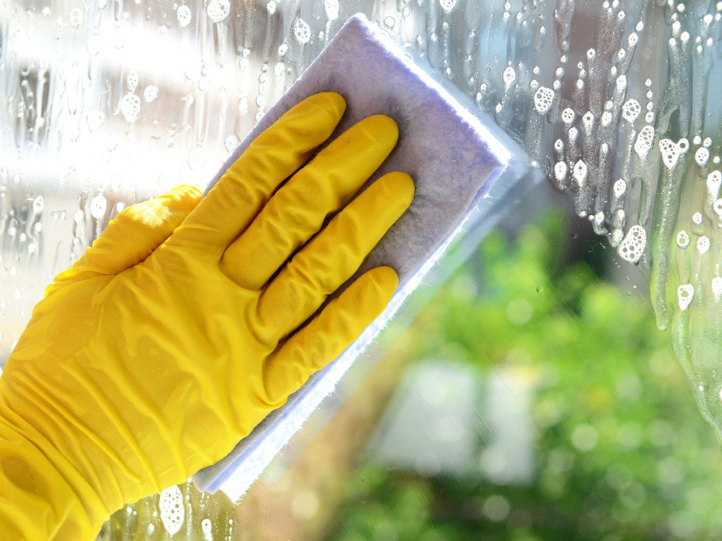 Yellow-gloved hand wiping a window with a sponge, cleaning water droplets from glass.