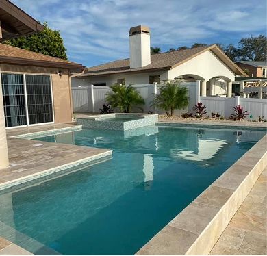 Pool area with turquoise water, beige tile, and a raised spa. A house is in the background.