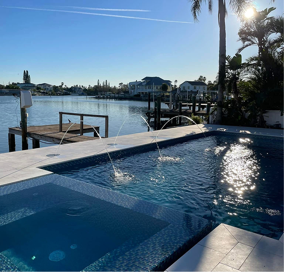 Pool with water fountains overlooking a waterway with houses and a dock on a sunny day.