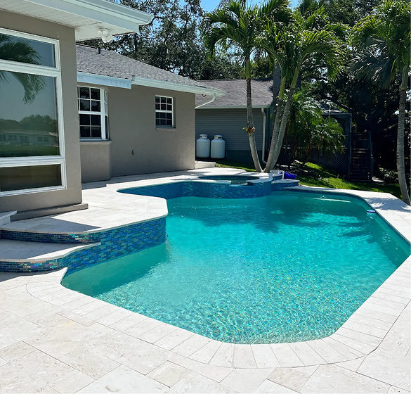 Backyard pool with turquoise water, surrounded by light-colored paving stones. Exterior of home and palm trees in background.