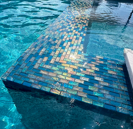 Pool steps with blue and iridescent mosaic tiles submerged in clear water.