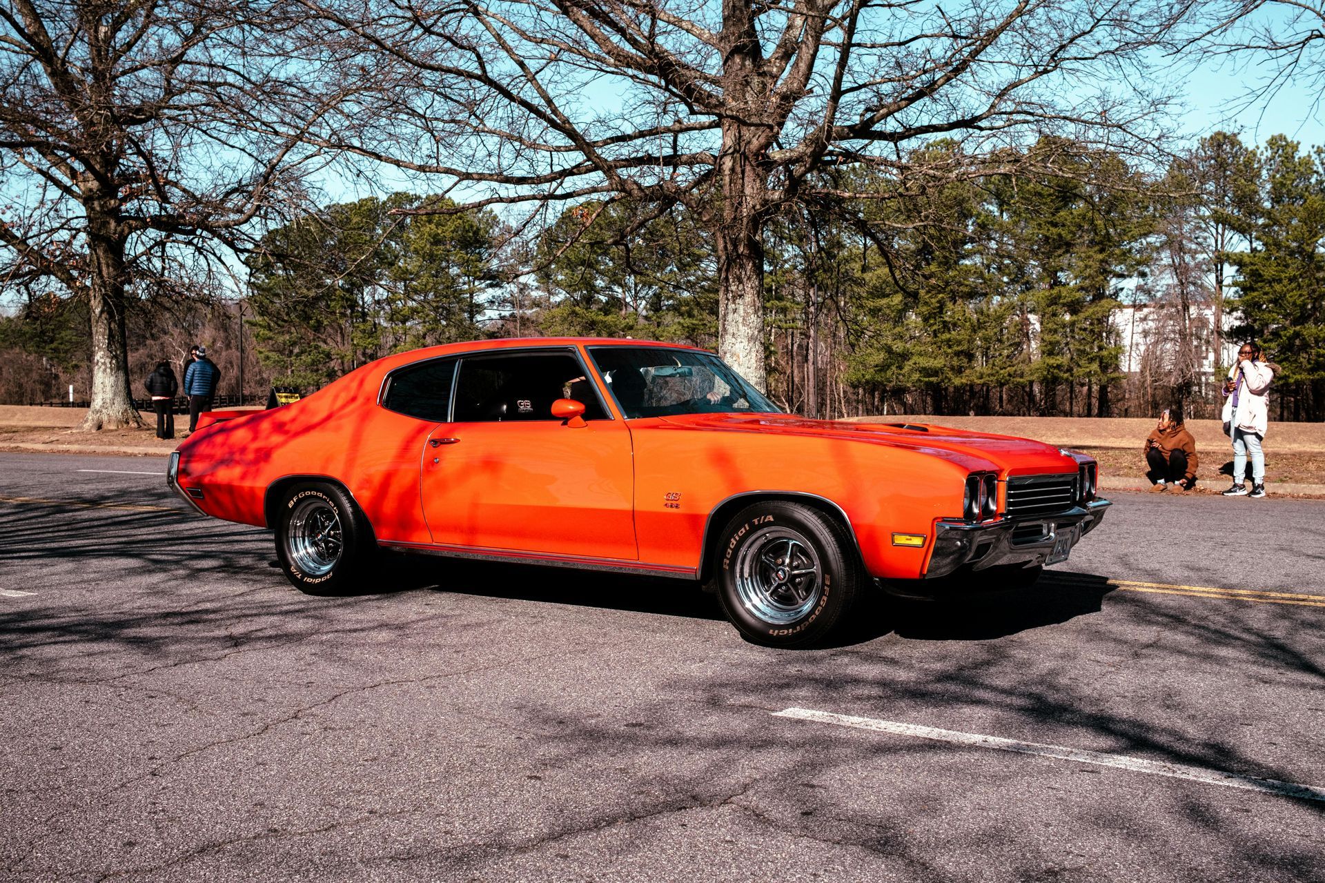 Bright orange classic muscle car parked on pavement in a park, with trees and people in the background
