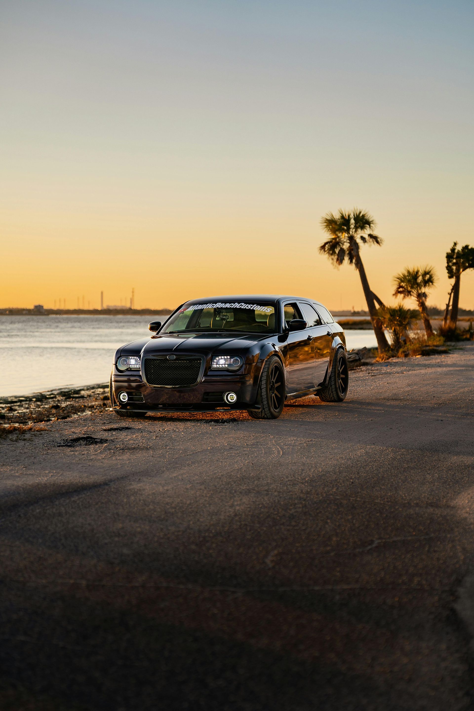 Black SUV parked by a waterfront at sunset with palm trees and calm water in the background