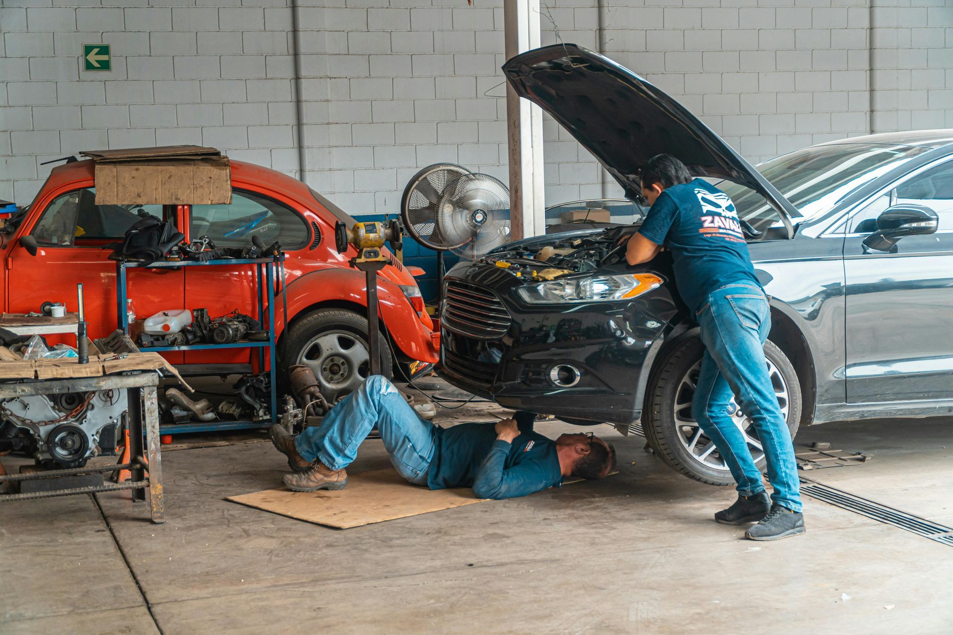 Mechanics repairing a silver SUV in a garage, with a red car and engine parts nearby