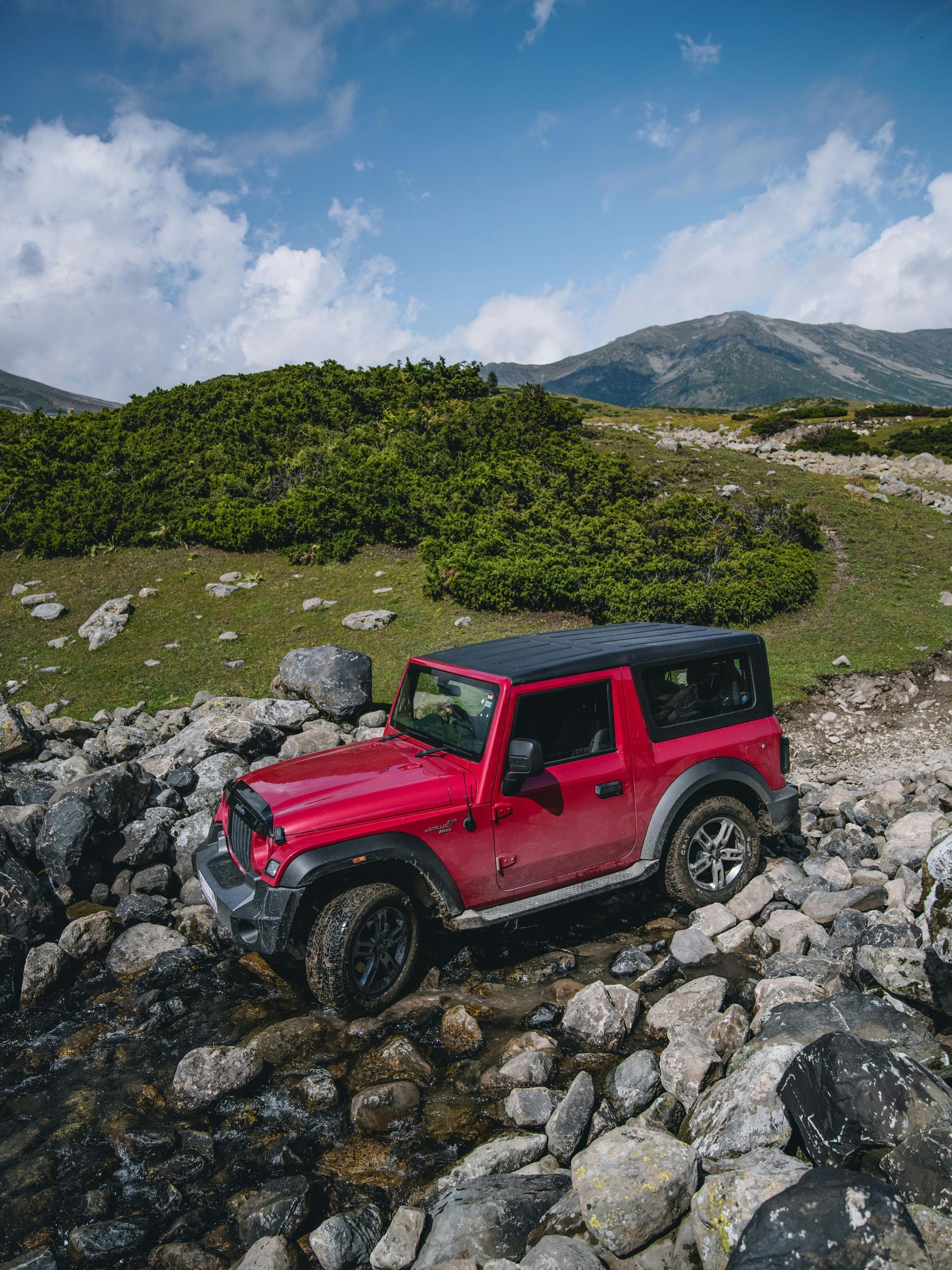 Red Jeep Wrangler climbing rocky terrain with mountains and cloudy sky in the background