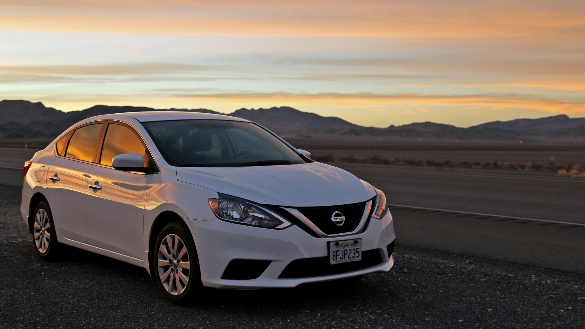 White sedan parked on a road at sunset with mountains in the background