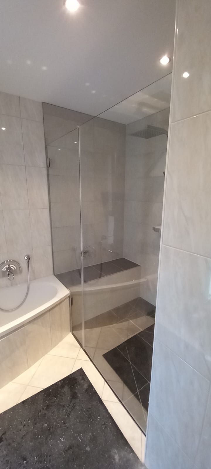 Doccia moderna in bagno con box in vetro, piastrelle grigie e tappetino scuro sul pavimento