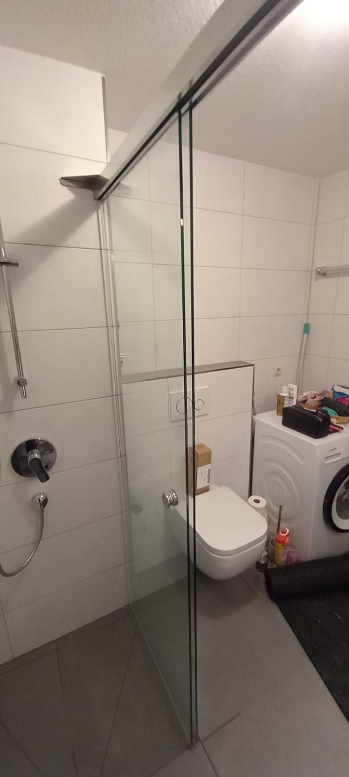 Piccolo bagno con pareti piastrellate di bianco, WC, doccia e lavatrice visibili dietro una parete divisoria in vetro.