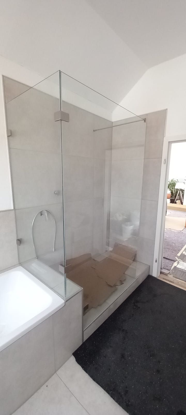 Doccia in bagno con box in vetro, vasca bianca, pavimento piastrellato color sabbia e tappetino scuro