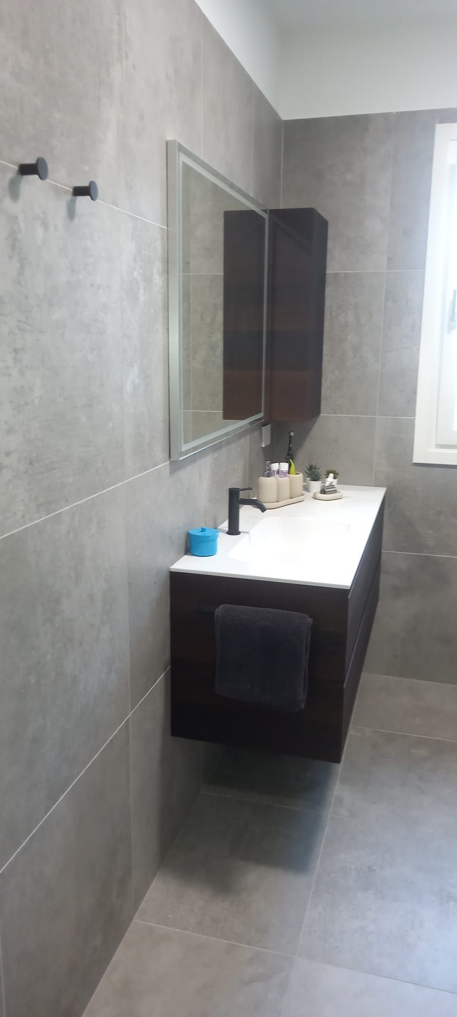 Bagno moderno con piastrelle grigie, mobile lavabo sospeso, armadietto a specchio e una finestra sulla destra.
