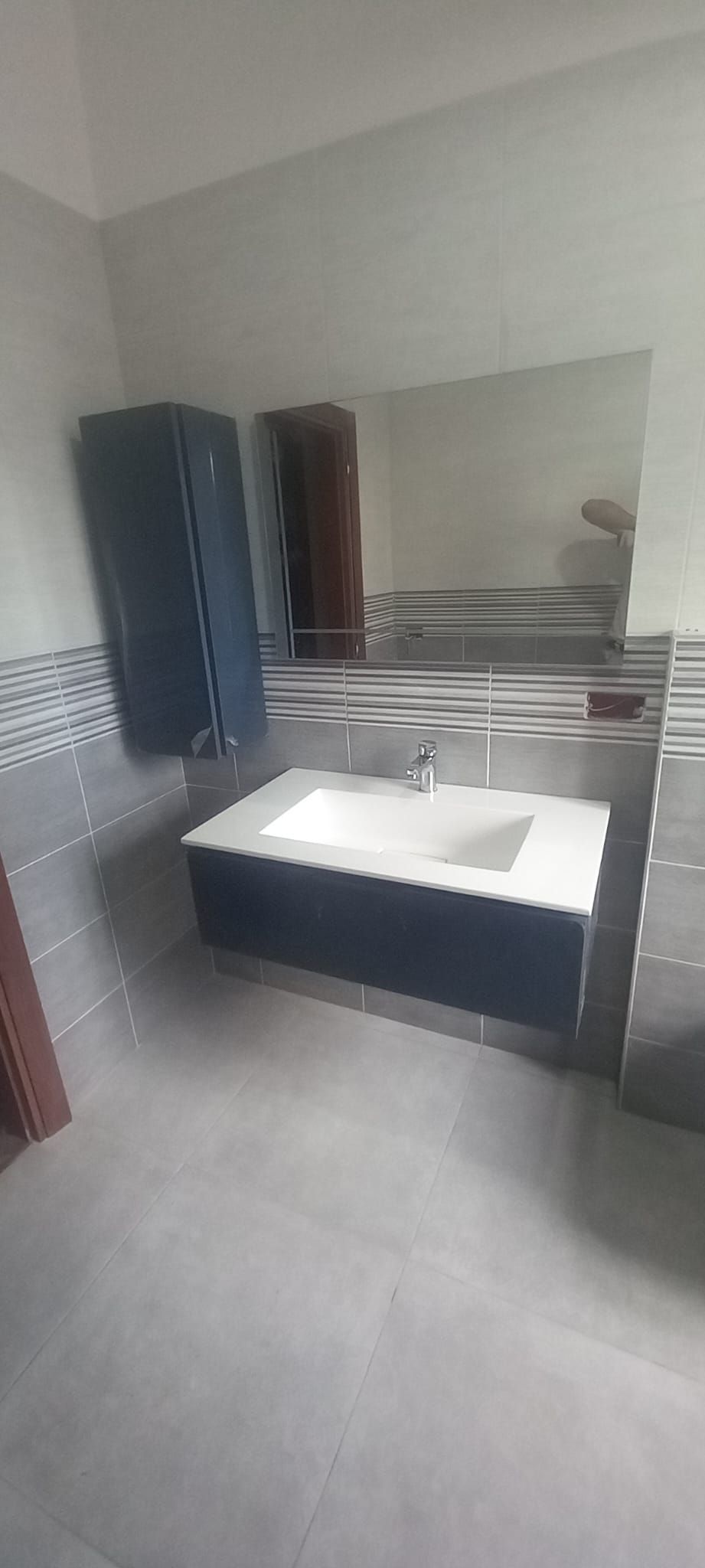 Bagno moderno con piastrelle grigie, un mobile lavabo bianco e un mobile blu scuro con pannello a specchio.