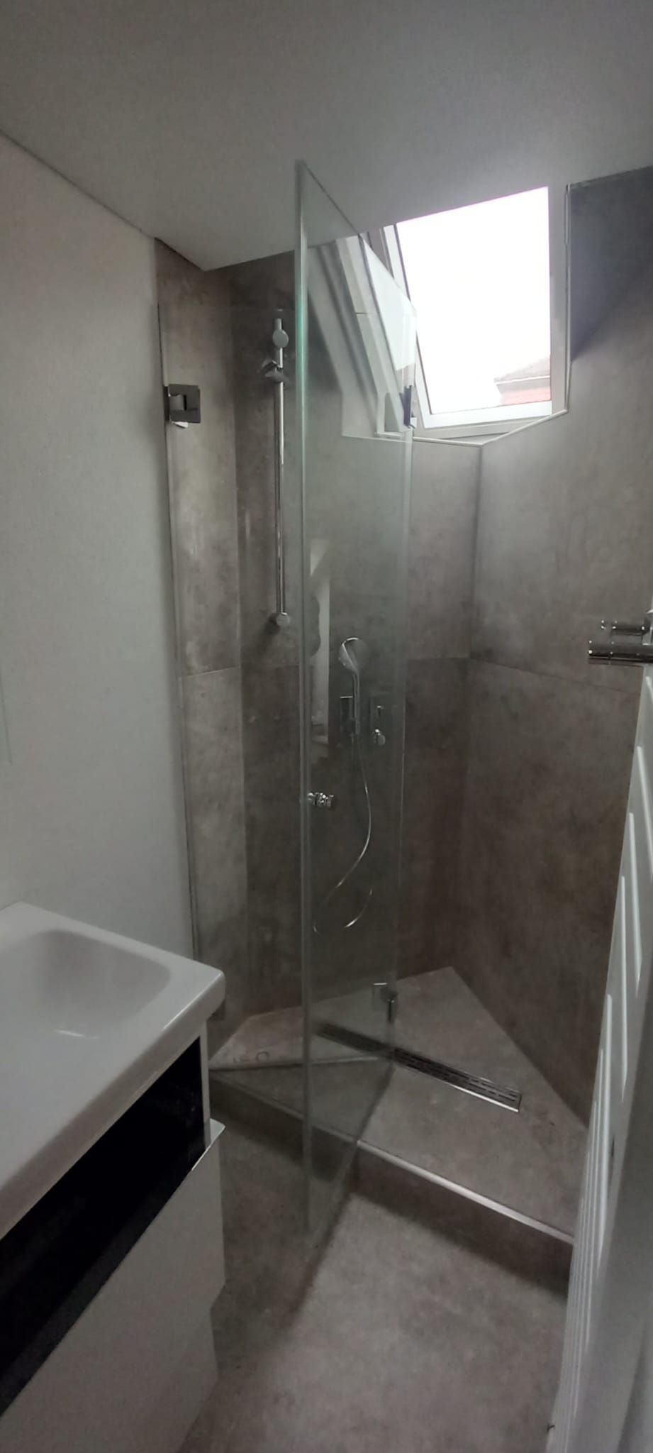 Piccolo bagno con box doccia in vetro, lavabo bianco e una piccola finestra sopra la doccia.