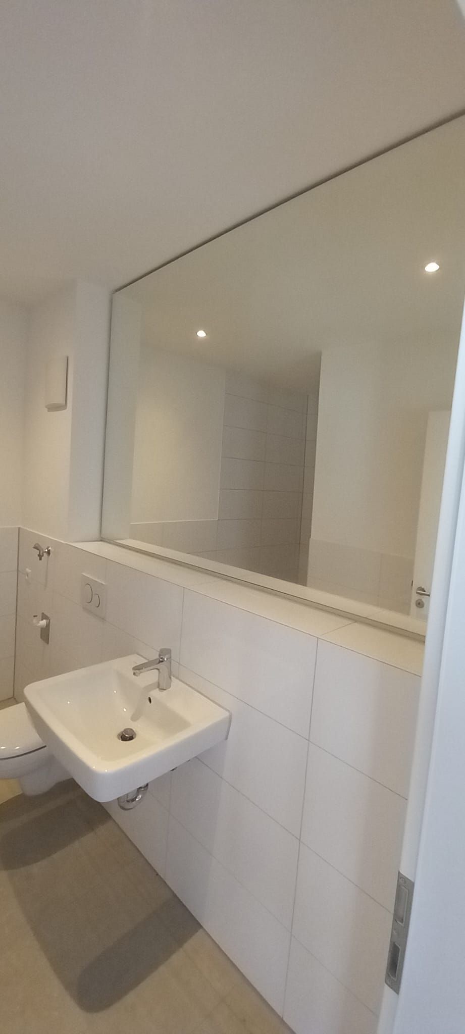 Bagno pubblico piastrellato di bianco, con lavabo a parete e un grande specchio sotto un soffitto inclinato.