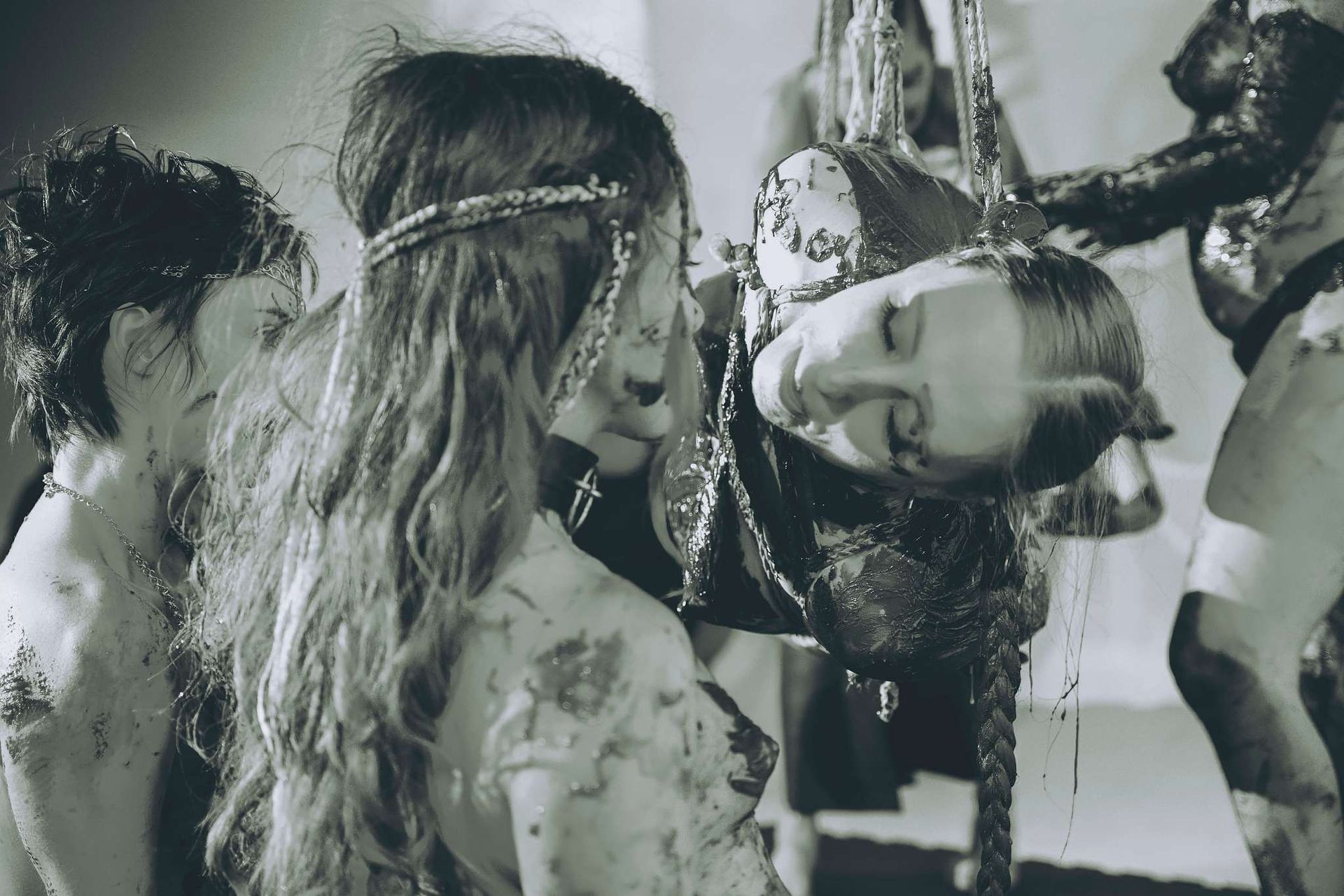 Black-and-white image of several figures covered in chocolate interacting with a woman suspended in rope.