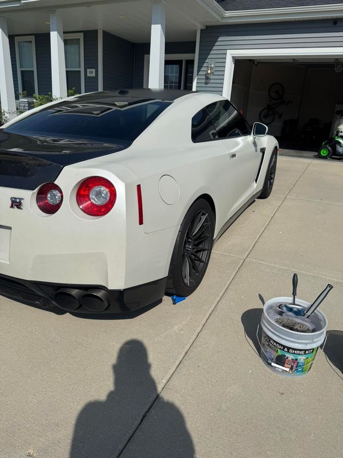 A white Nissan GT-R parked in a driveway next to a bucket of cleaning supplies, with a person's shadow in the foreground.