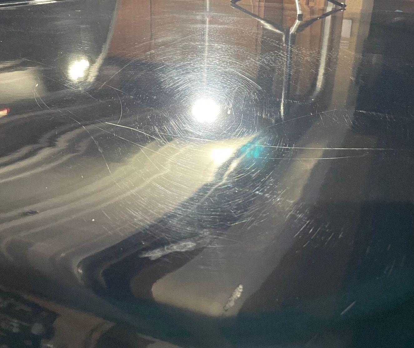 Close-up of a shiny black surface with many scratches and light reflections, likely a car hood.