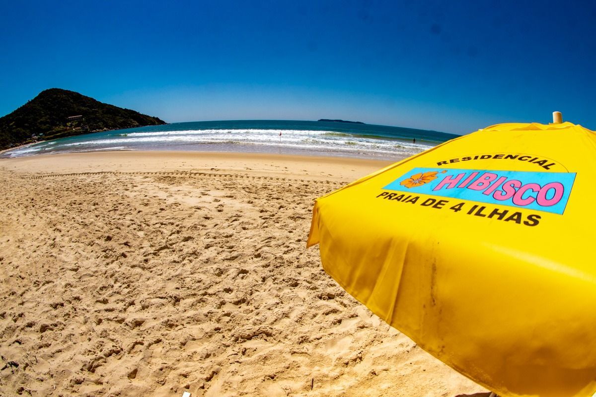 Praia ensolarada com oceano azul, areia e um guarda-sol amarelo em primeiro plano