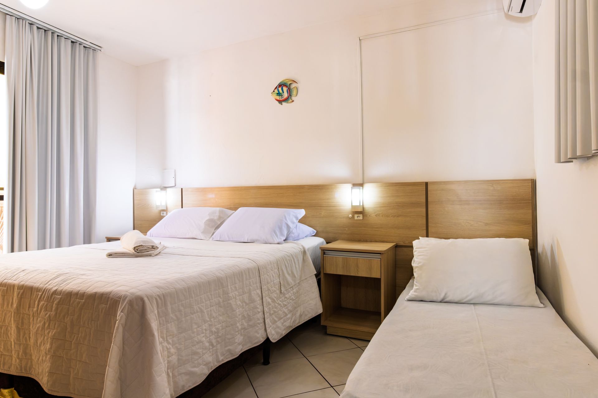 Quarto de hotel com duas camas, cabeceira de madeira, luminárias de cabeceira e cortinas, em um ambiente claro em tons de bege