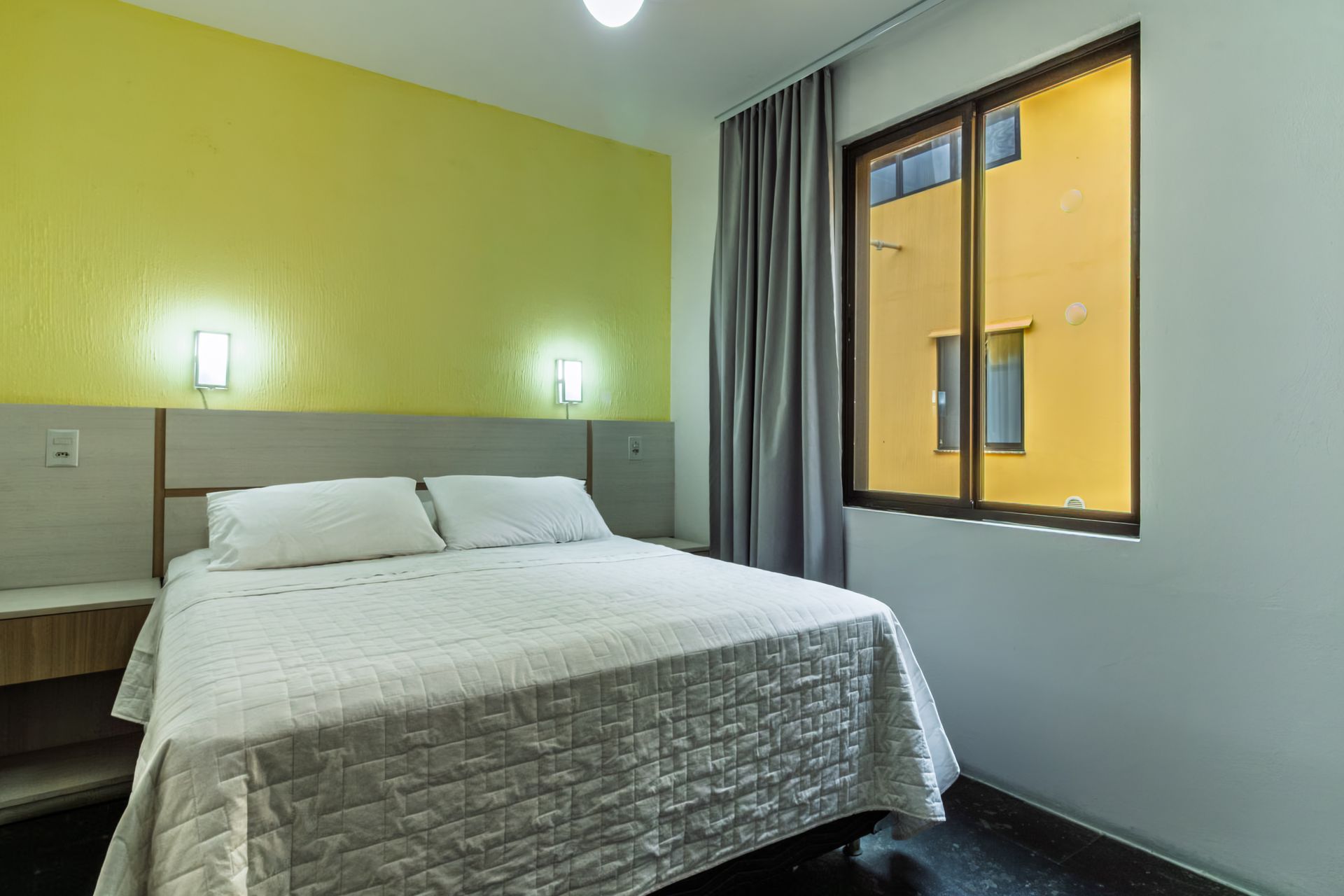 Quarto moderno com cama branca, parede de destaque verde, arandelas e uma janela com cortinas