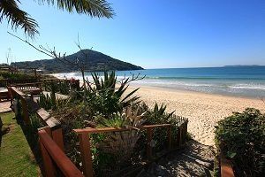 Praia com águas azuis e cristalinas, areia, cerca de madeira e uma colina ao longe sob um céu ensolarado.