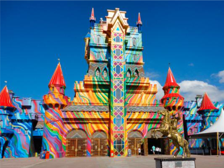 Edifício colorido que lembra um castelo, com padrões vibrantes em arco-íris e torres vermelhas, sob um céu azul límpido