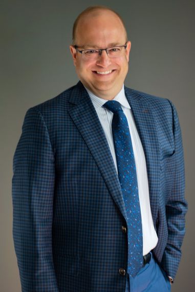 Smiling man in a blue suit and tie, standing against a gray studio backdrop