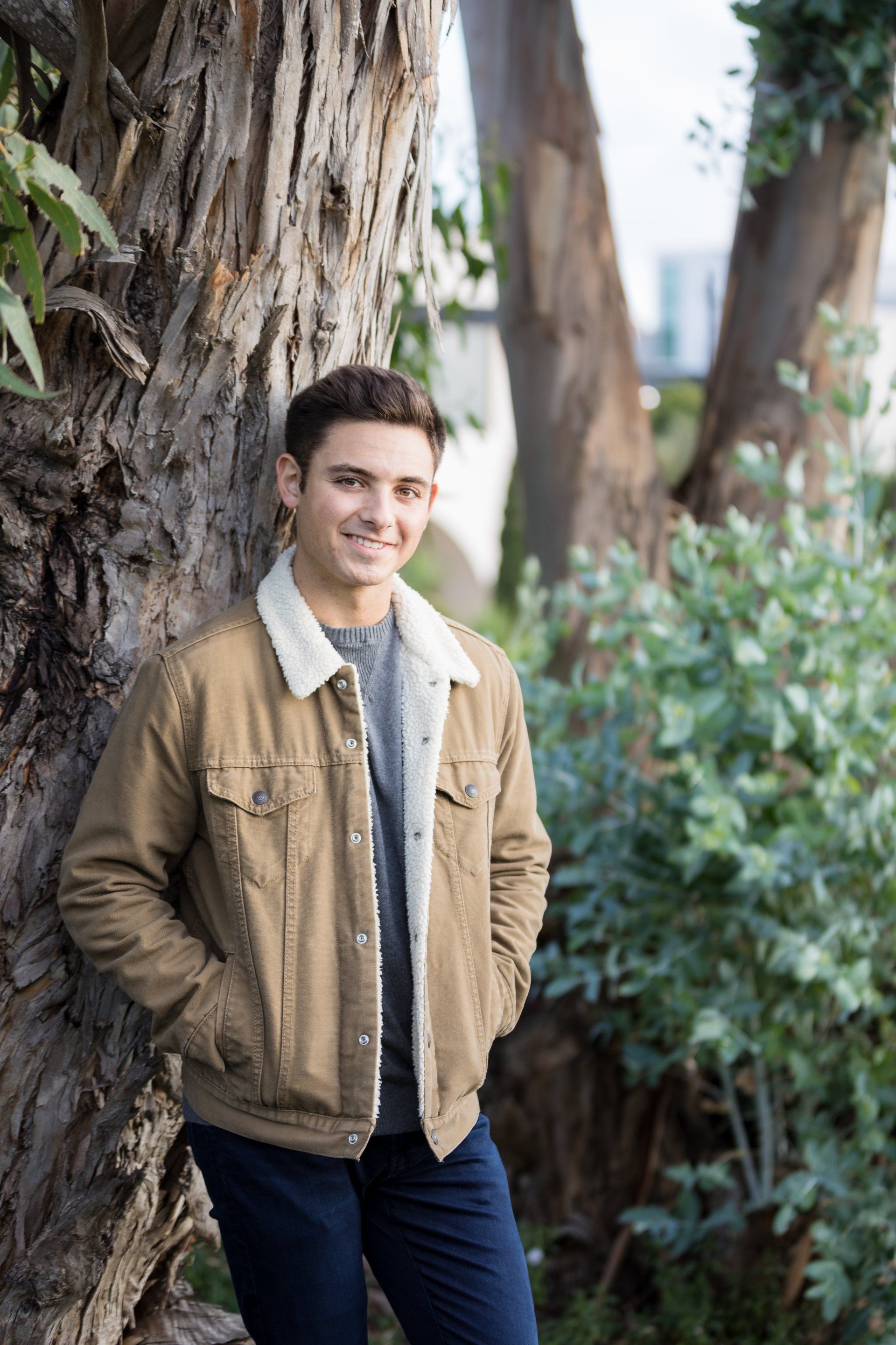 A young man in a brown jacket is leaning against a tree.