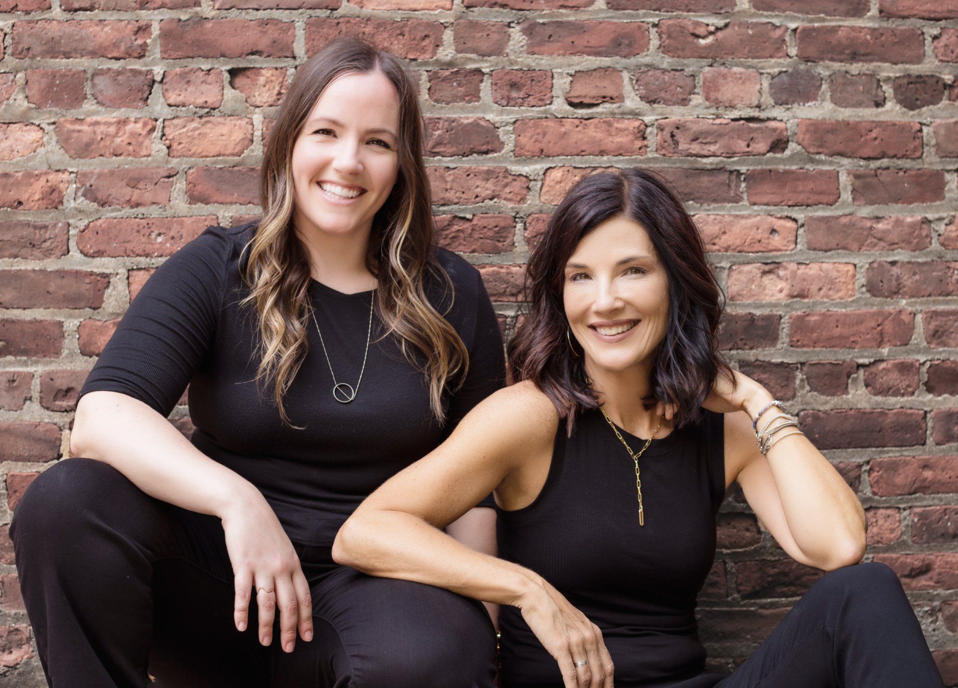 Two women are sitting next to each other in front of a brick wall.