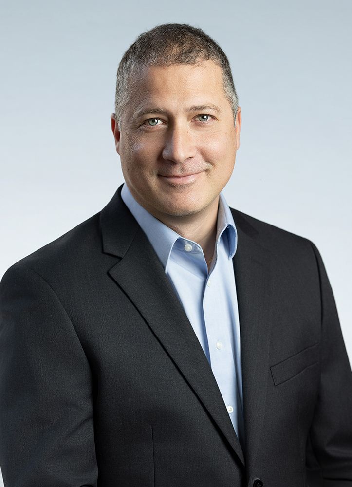 Studio portrait of a smiling man in a dark suit and light blue shirt on a gray background